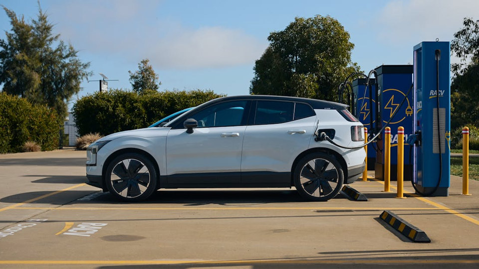A white electric vehicle is plugged into a charging station at an outdoor parking lot, with trees and a partly cloudy sky in the background.